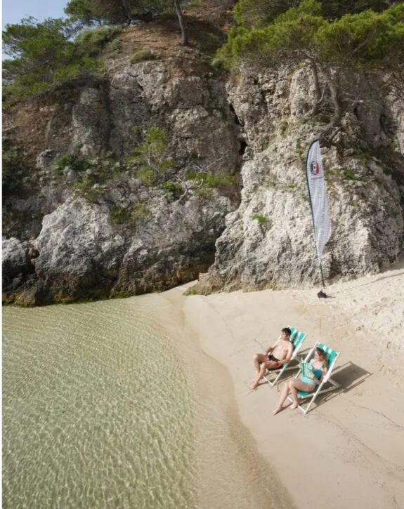 Due persone si rilassano su sdraio sulla spiaggia vicino a una scogliera, con acqua limpida e sabbia dorata.