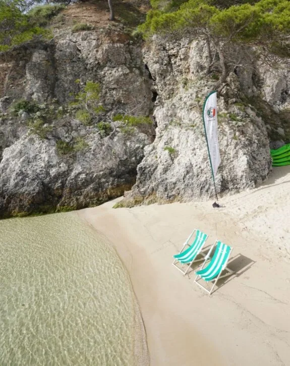 Due sedie a sdraio sul mare vicino a scogli e vegetazione, con bandiera pubblicitaria sulla spiaggia.