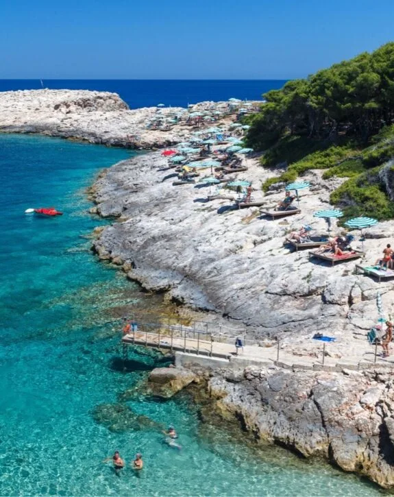 Spiaggia rocciosa con lettini, ombrelloni e persone che nuotano e prendono il sole, con il mare cristallino e il cielo sereno.