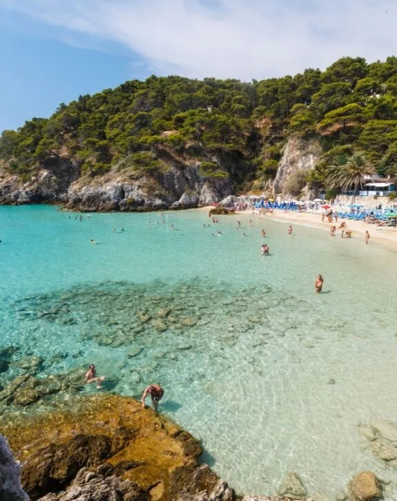 Spiaggia con acque cristalline, rocce, ombrelloni e persone che nuotano e si rilassano. Collina verde sullo sfondo.