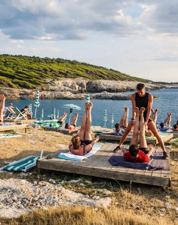 Persone che praticano yoga sulla spiaggia di fronte al mare con ombrelloni e scogliere sullo sfondo.
