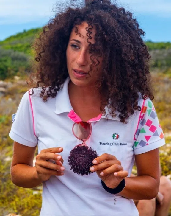 Donna con capelli ricci, indossa una maglietta del Touring Club Italiano e tiene un riccio di mare in mano, in un paesaggio naturale.