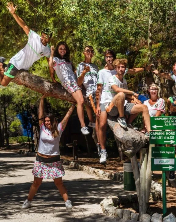 Gruppo di giovani che si diverte su un tronco d'albero in un parco, circondato da alberi e segnali stradali.