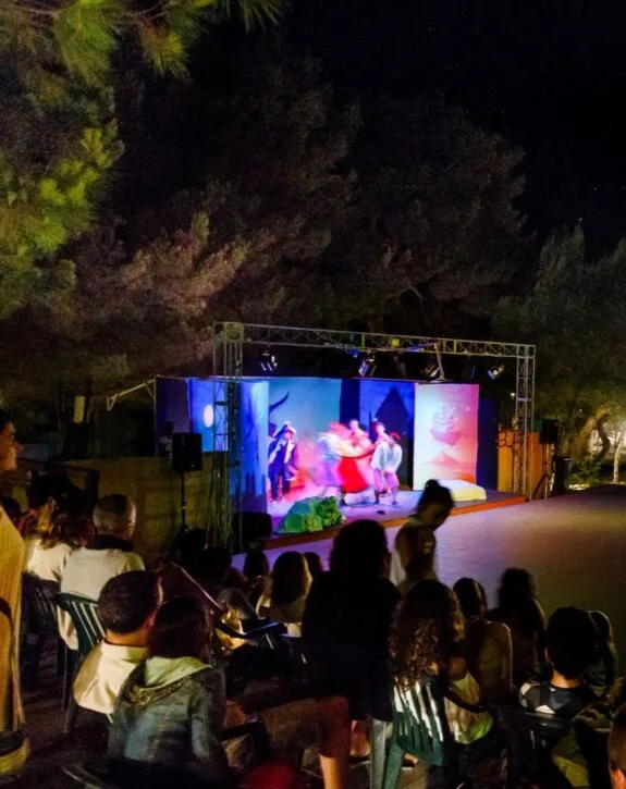 Spettacolo teatrale all'aperto con attori in abiti colorati, pubblico seduto e alberi sullo sfondo di notte.
