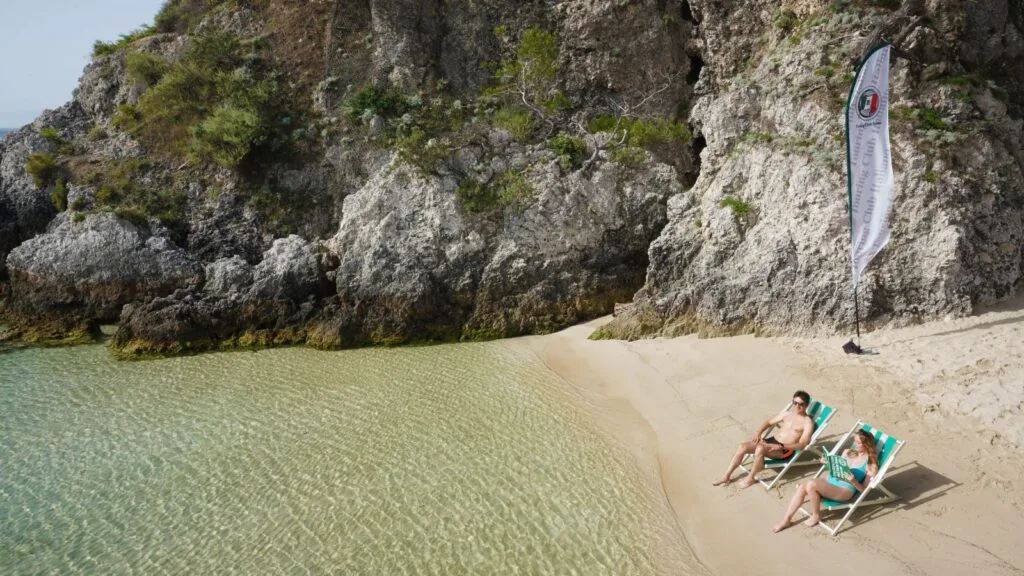 Due persone che si rilassano su sedie a sdraio sulla spiaggia di fronte a scogli e acqua cristallina, con bandiera della Club.