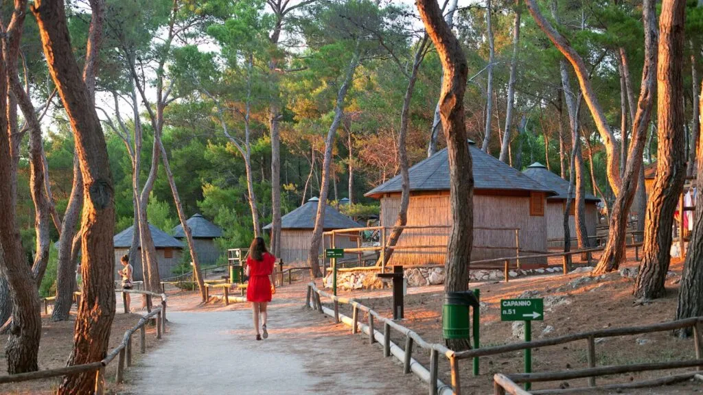 Donna in abito rosso cammina tra le capanne in un bosco illuminato dal tramonto.