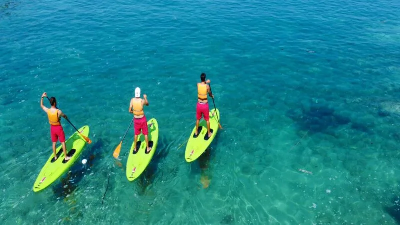 Tre persone in paddleboard con superfici verdi, indossano giubbotti gialli e pantaloncini rosa, sul mare azzurro.
