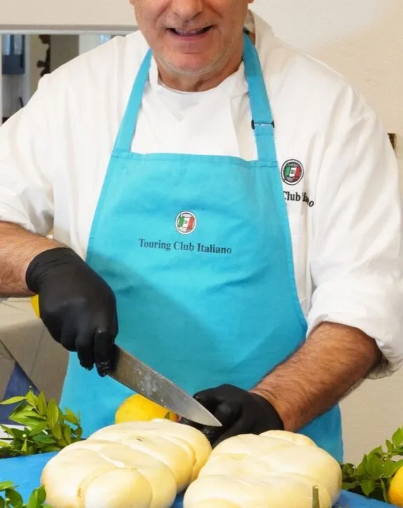 Chef sorridente in grembiule azzurro, mentre affetta formaggio fresco su un tavolo decorato con limoni e foglie verdi.