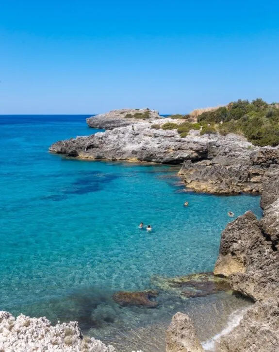 Acqua cristallina in una baia rocciosa, con persone che nuotano e un cielo sereno sullo sfondo.