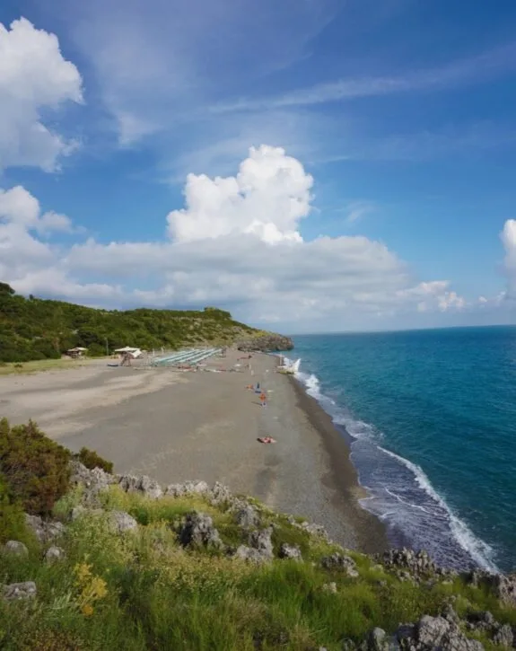 Spiaggia di ciottoli con ombrelloni e persone in riva al mare, incastonata tra colline verdi e un cielo blu.