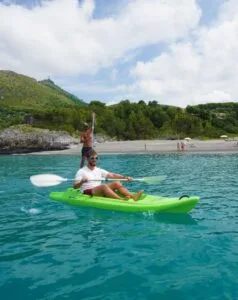 Due uomini in kayak verde navigano in acque turchesi, con una spiaggia e colline verdi sullo sfondo.