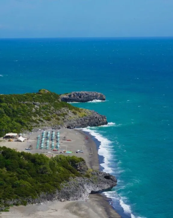 Esperienza panoramica di una spiaggia con ombrelloni e onde che si infrangono su una costa rocciosa e verde.