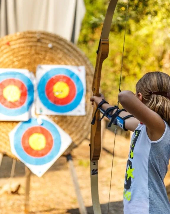 Ragazza che tira con l'arco verso tre bersagli colorati su uno sfondo naturale.