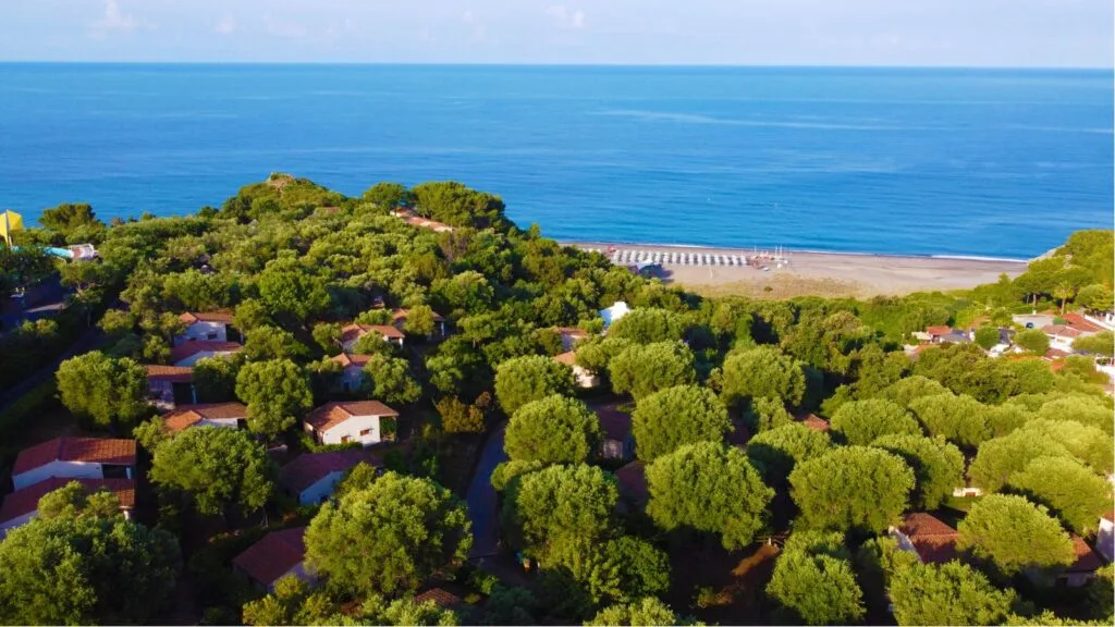 Vista aerea di case circondate da alberi e una spiaggia con ombrelloni e il mare all'orizzonte.