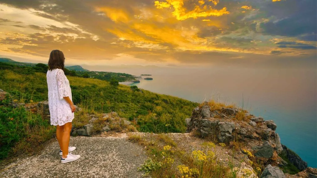 Donna in abito bianco osserva il tramonto su una scogliera con vista sull'oceano e paesaggi verdi.