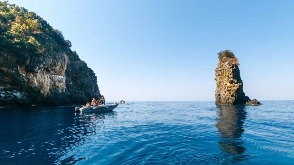 Barca con persone vicino a una scogliera e a unisolotto in mare aperto, sotto un cielo sereno.