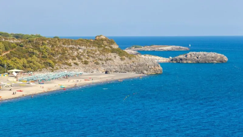 Spiaggia con lettini e ombrelloni vicino a scogliere e acque blu, sullo sfondo il mare e il cielo.