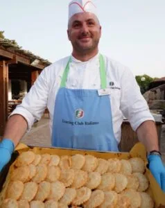 Un cuoco sorride mentre tiene un vassoio di dolci fritti, indossando un grembiule azzurro e un cappello da chef.