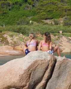 Due ragazze sedute su una grande roccia, osservano il mare e il paesaggio verdeggiante dietro di loro.