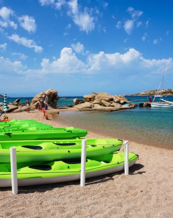 Città di spiaggia con canoe verdi sulla sabbia, acque cristalline e rocce, sotto un cielo blu con nuvole bianche.