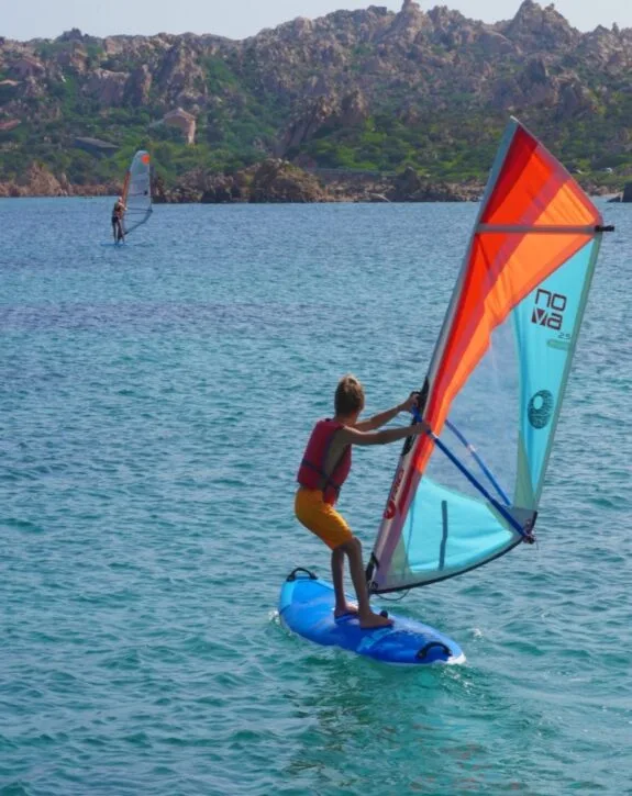 Due persone praticano windsurf in un mare blu, con rocce e vegetazione verde sullo sfondo.