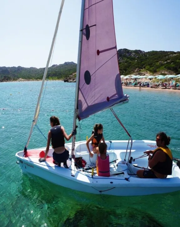 Gruppo di quattro persone su una barca a vela in acqua cristallina, con una spiaggia e colline sullo sfondo.
