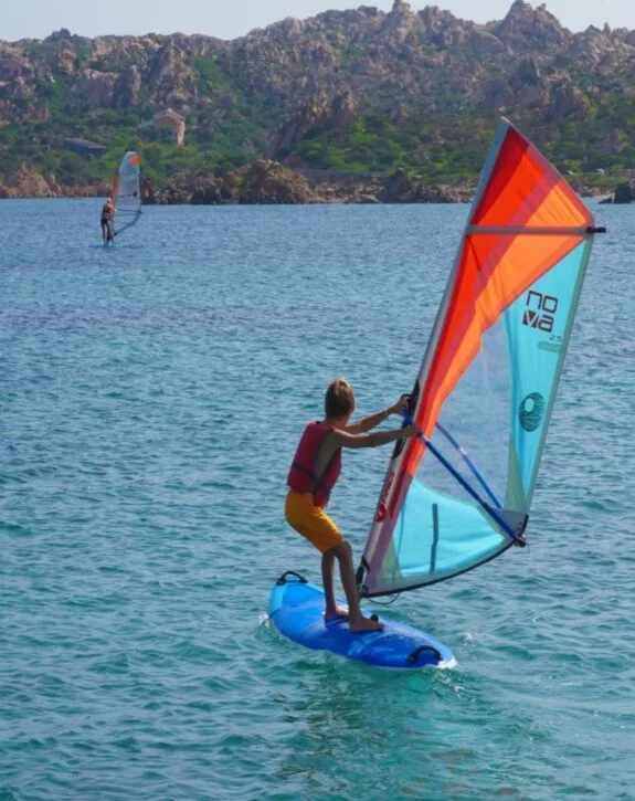 Giovane in pratica di windsurf su acqua turchese, circondato da montagne. Vento che gonfia la vela colorata.