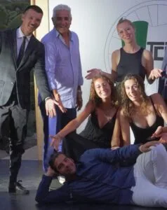 Gruppo di sei persone sorridenti in abiti eleganti su un palco, con uno sdraiato a terra e gli altri in posizioni gioiose.