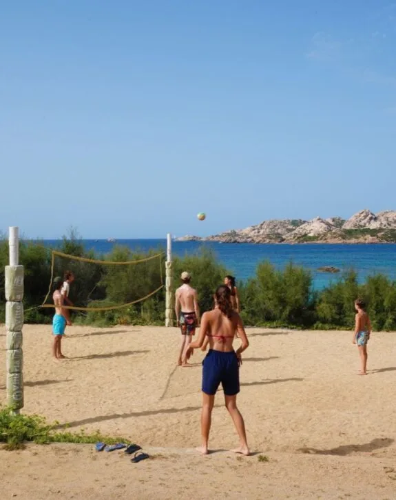 Giovani giocano a beach volley su una spiaggia sabbiosa, con un mare azzurro e colline rocciose sullo sfondo.