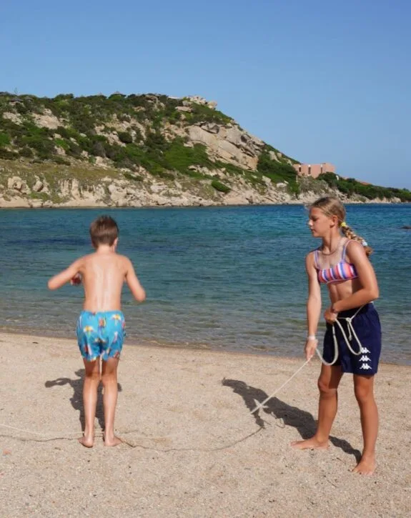 Due bambini giocano sulla spiaggia: una ragazza tiene una corda mentre un ragazzo salta vicino all'acqua blu.