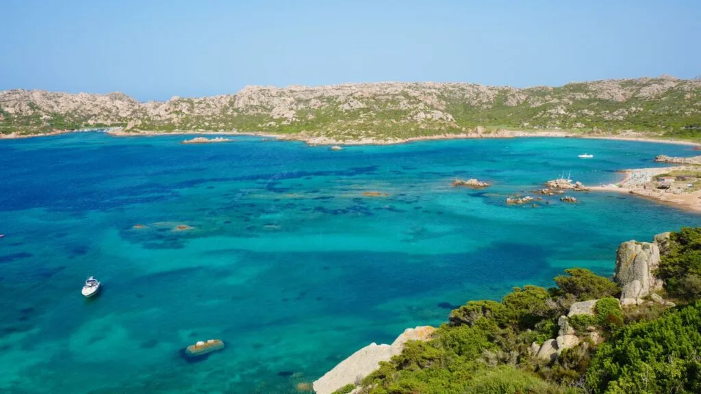 Vista di una baia con acque cristalline e scogli, sfondo di colline verdi e cielo azzurro.