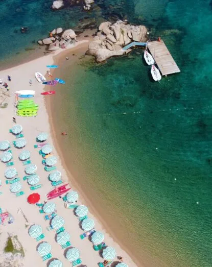Spiaggia con ombrelloni, kayak e barche ancorate in acque cristalline, vicino a grandi rocce e un pontile in legno.