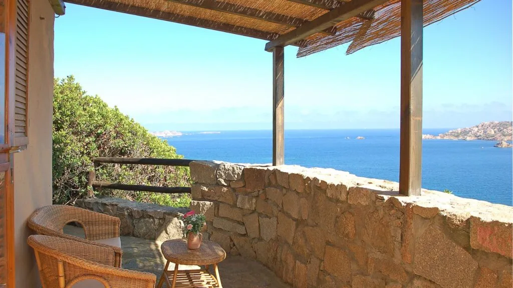 Balcone in pietra con vista sul mare, sedie di vimini e tavolino con vasi di fiori, sotto un pergolato di legno.
