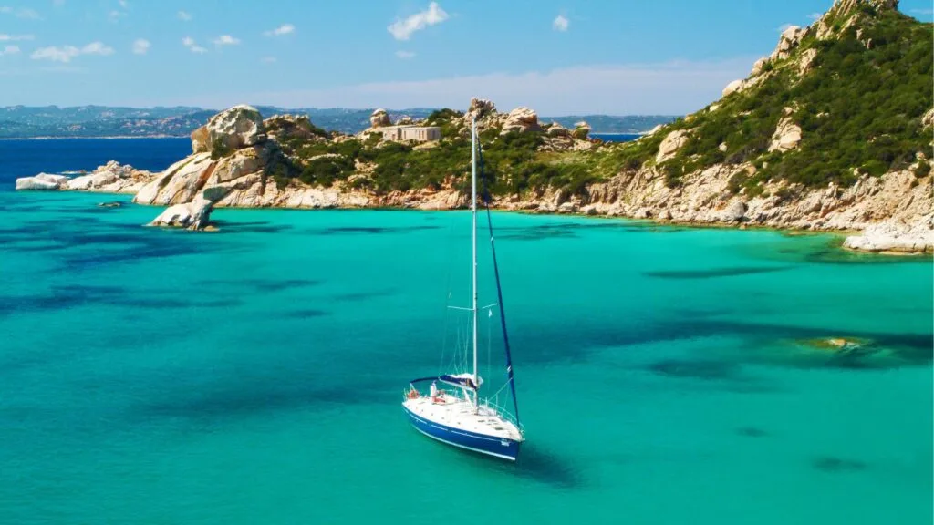 Moltissimo mare azzurro con barca a vela e scogliera verde sullo sfondo.