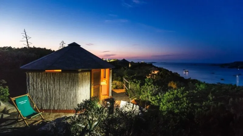Casa di legno con vista sul mare al tramonto, illuminata tra la vegetazione.