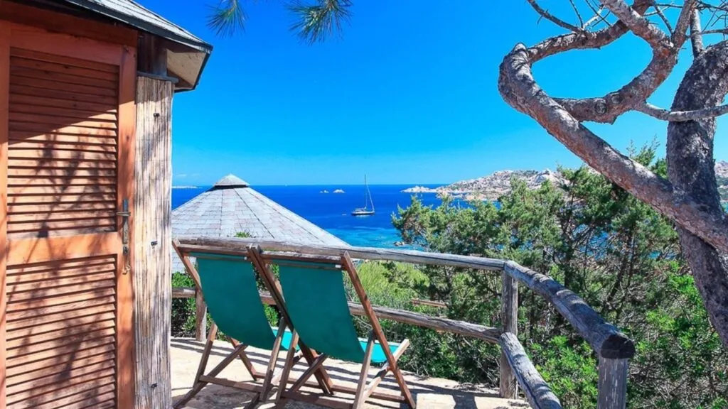 Balcone con sedie e tetto di paglia, vista sul mare e su una barca in acque cristalline, circondato da vegetazione e cielo azzurro.