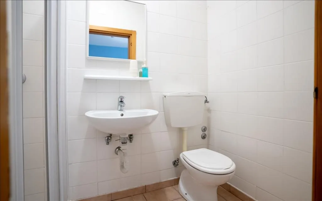 Bagno con lavabo, WC, specchio e piccola finestra, pareti in piastrelle bianche