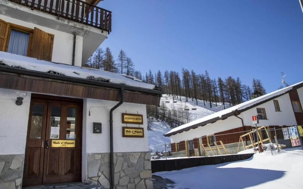 Ingresso del Residence Hotel Nube d'Argento con insegna in legno, neve e seggiovia sullo sfondo