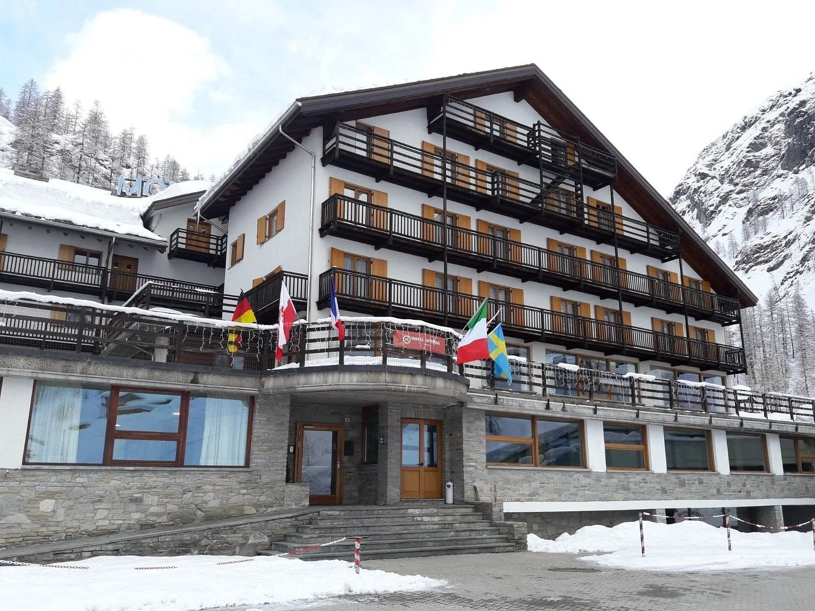 Facciata dell'hotel con bandiere internazionali, ingresso in pietra e balconi in legno sotto la neve