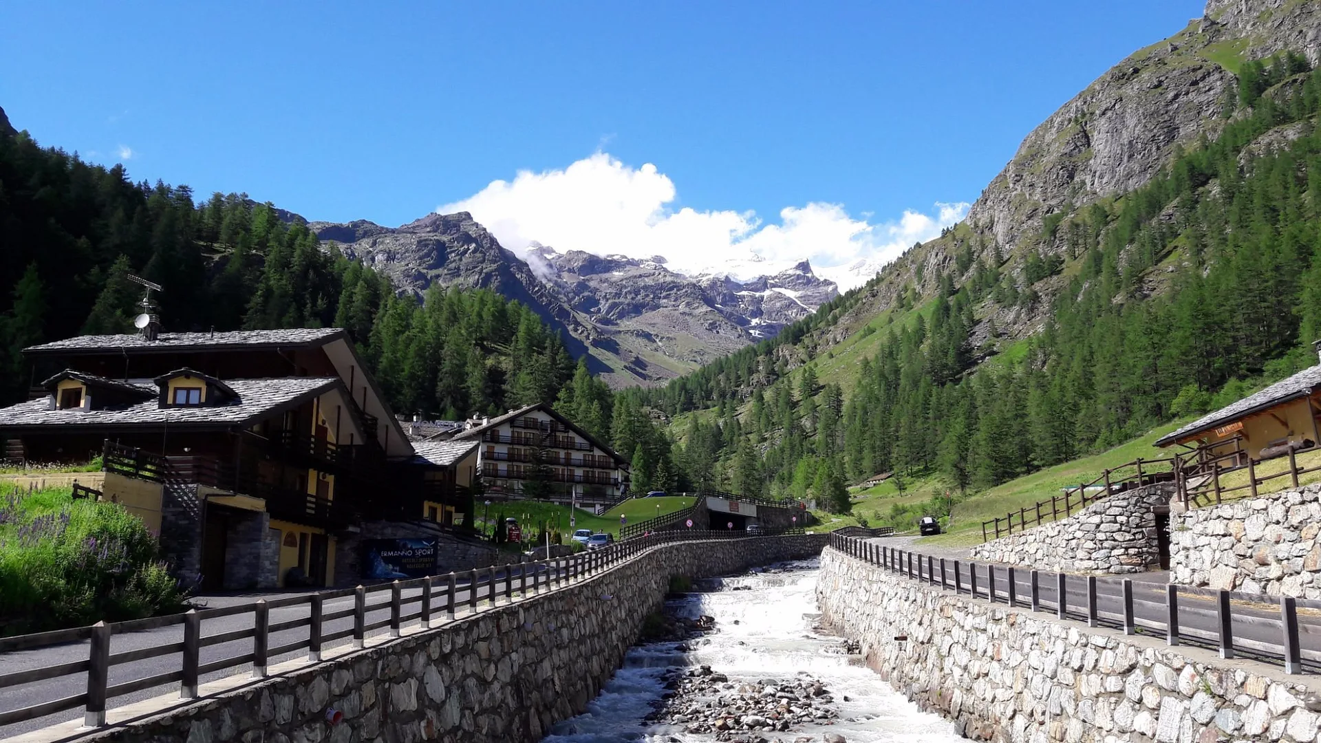 Veduta estiva della valle con torrente, strada di montagna e prati verdi tra le vette alpine