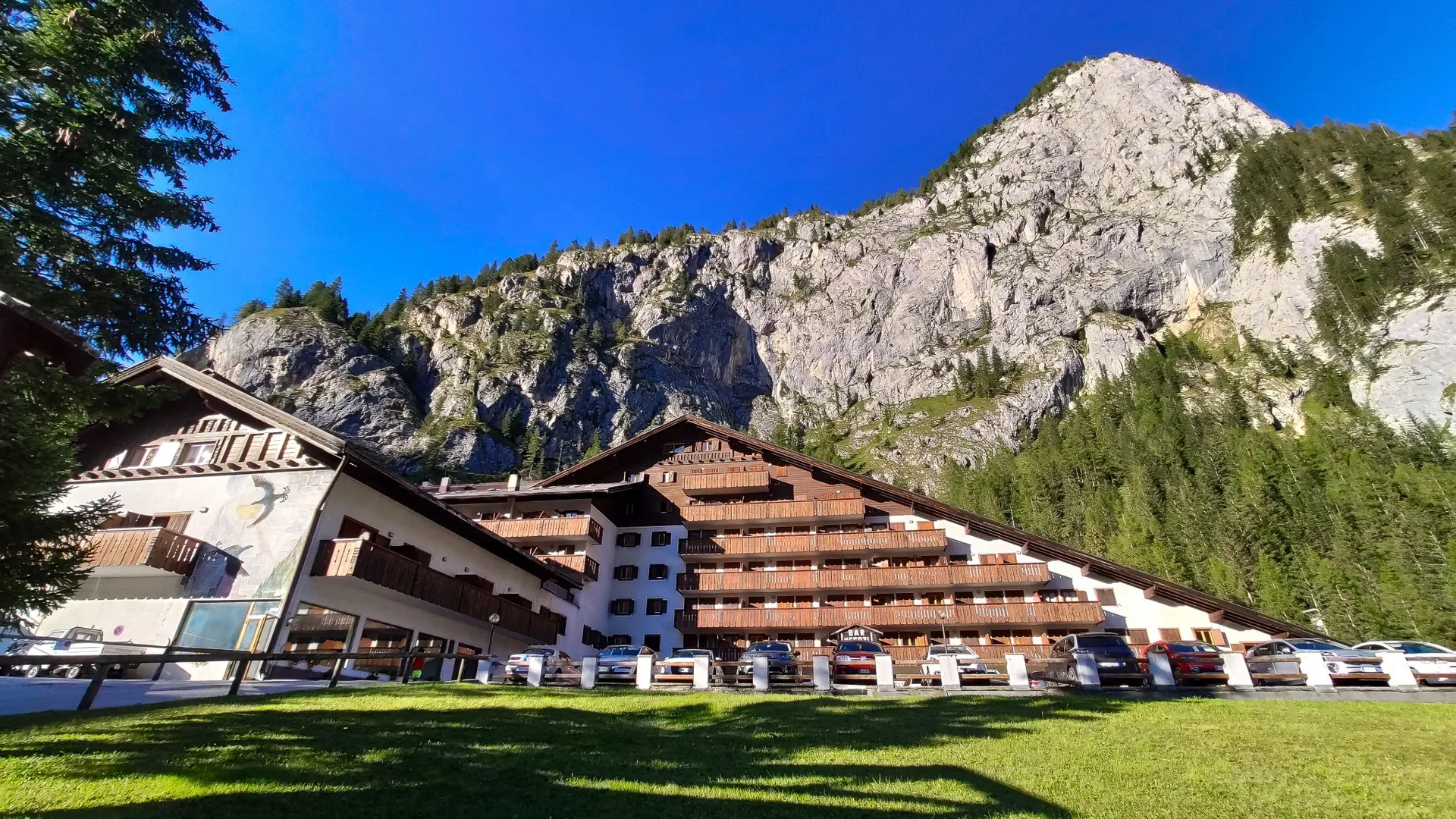Esterno dell'hotel in estate con prato verde, parcheggio e montagna rocciosa sullo sfondo