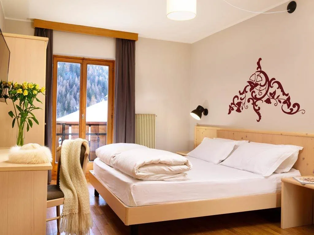 Camera luminosa con letto matrimoniale, decoro floreale a parete e finestra sul paesaggio montano