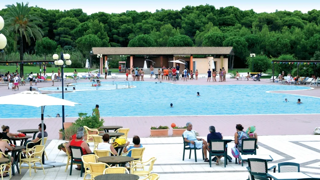 Piscina con area solarium, tavoli e ombrelloni circondata da pini