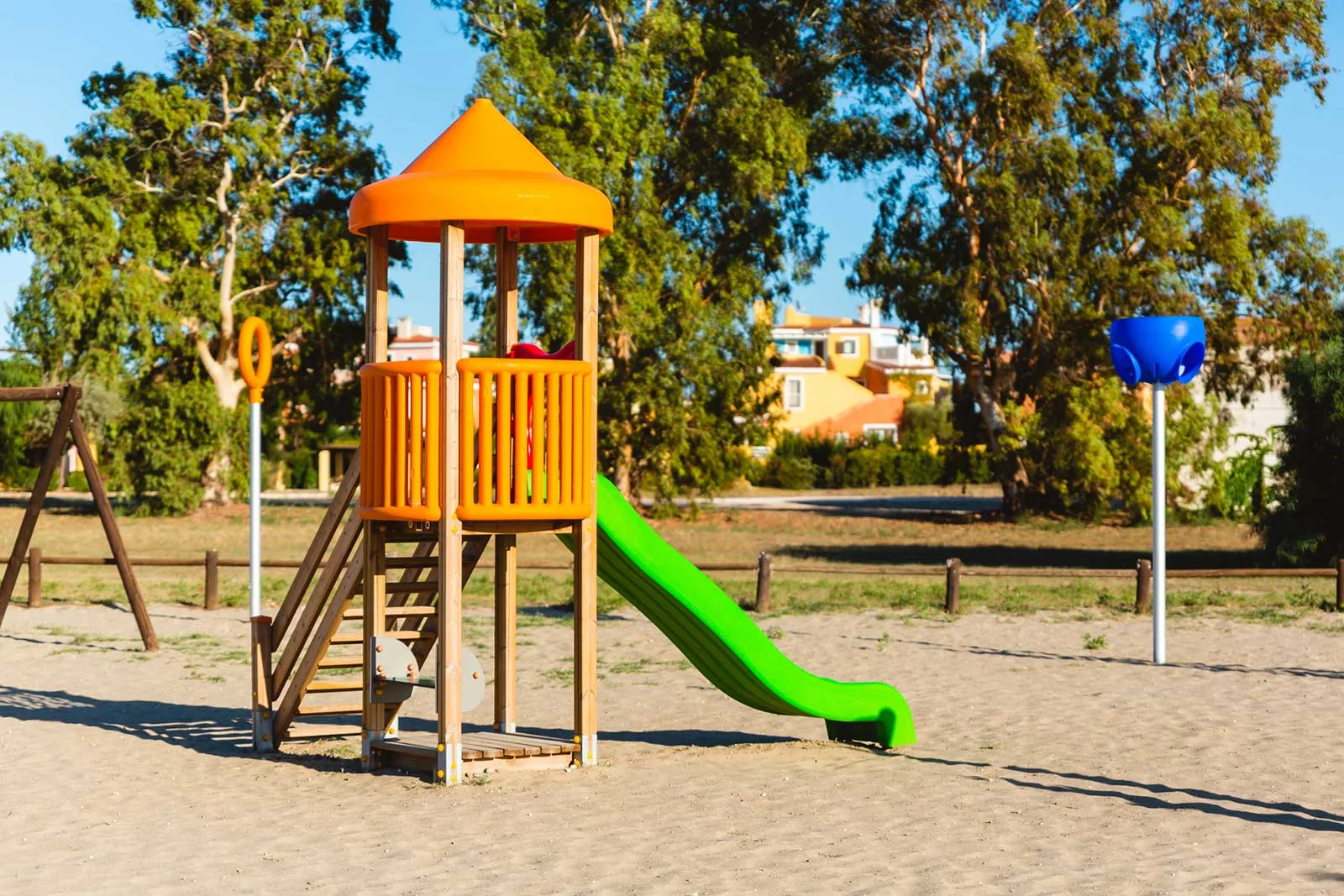 Area giochi per bambini con scivolo verde e struttura arancione su sabbia tra gli alberi