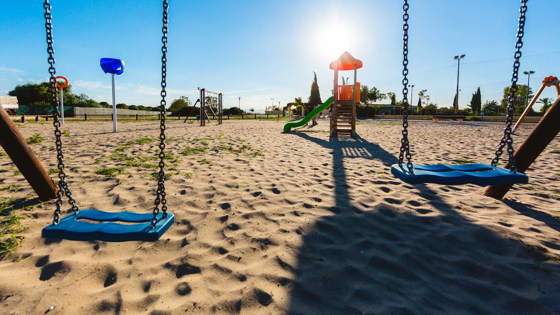 Parco giochi con altalene e scivolo su sabbia illuminato dal sole