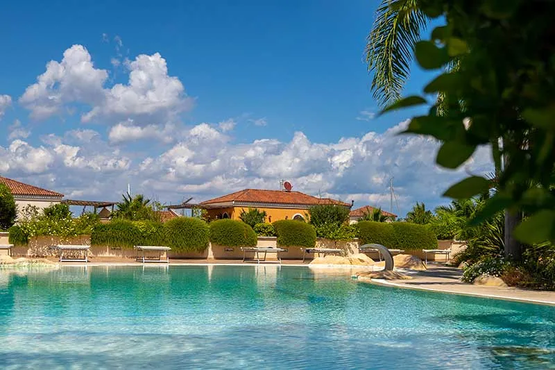 Piscina esterna con acqua turchese circondata da siepi verdi ed edifici sotto cielo nuvoloso