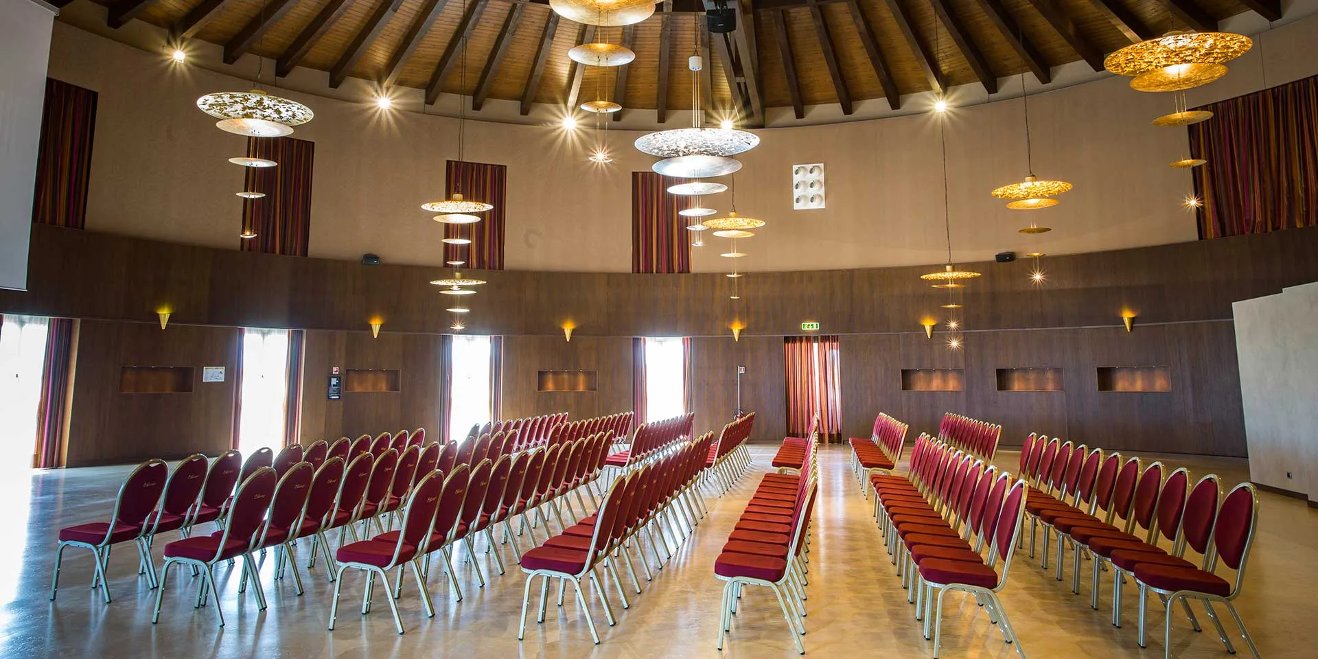 Sala meeting circolare con sedie rosse disposte a file, soffitto in legno e lampadari di design