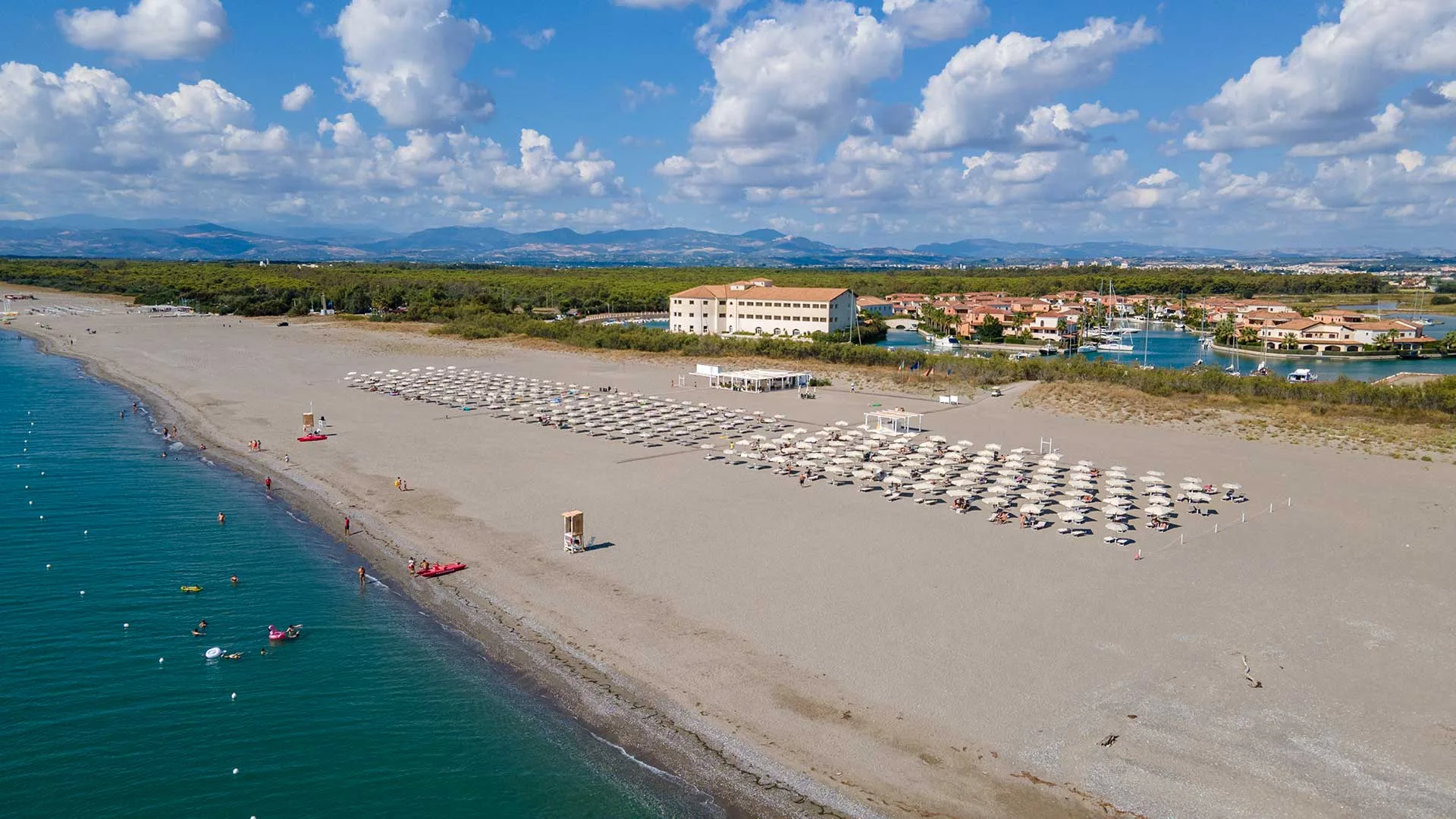 Panorama aereo della spiaggia sabbiosa con ombrelloni, mare e resort con porto sullo sfondo
