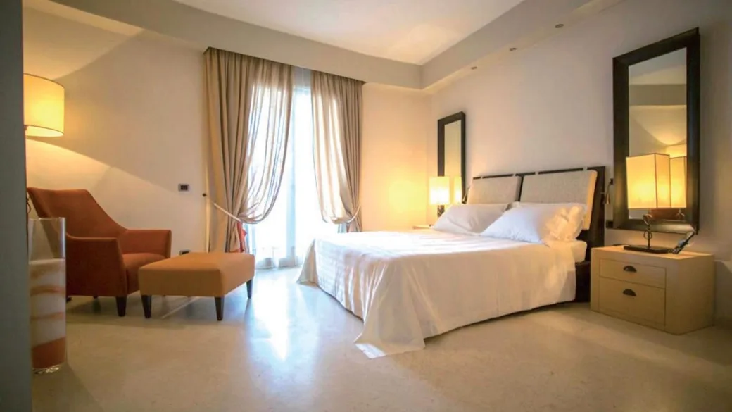 Camera matrimoniale elegante con letto, poltrona arancione, ampia finestra con tende e arredi moderni
