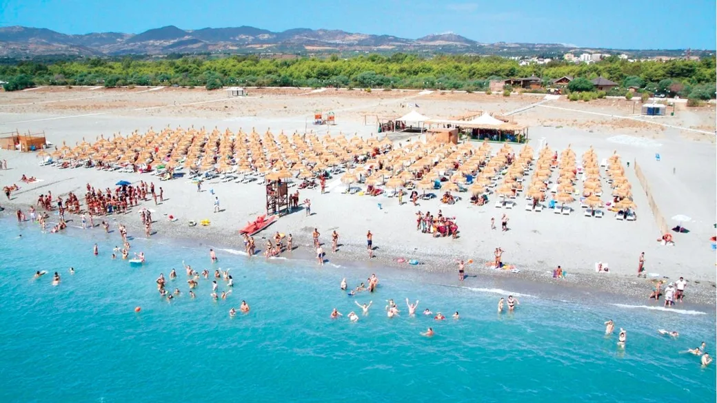 Spiaggia affollata con file di ombrelloni e bagnanti in acqua, colline sullo sfondo